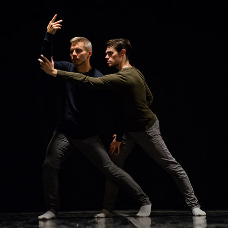 Two dance artists pose in a black box theatre. They both have one hand extended