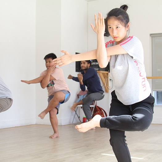 Students stand with their left legs bent onto their right knees. Their left arms are being stretch across their chests. They are in a dance class in a bright dance studio.