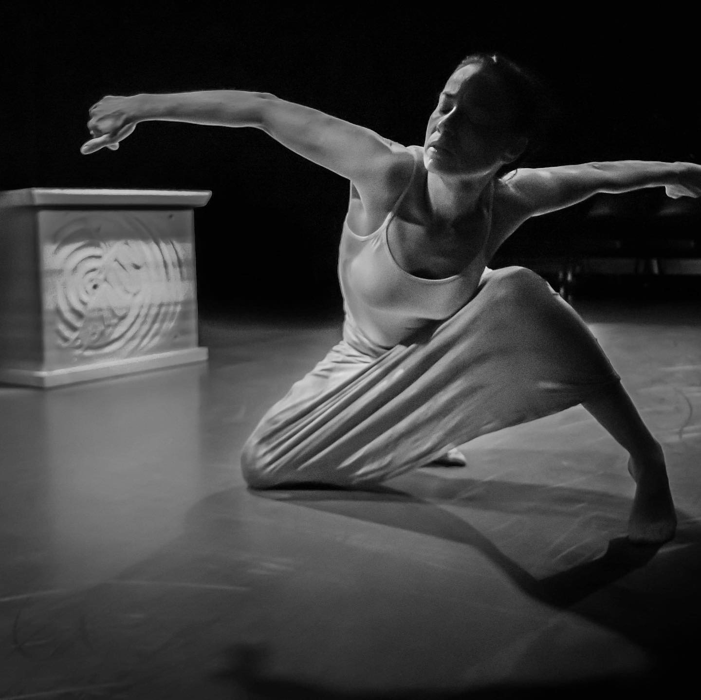Dance artist Tasha Faye Evans poses mid performance. Her arms are outstretched and she is kneeling down with her eyes closed. There is a large carved box behind her. This image is in black and white.