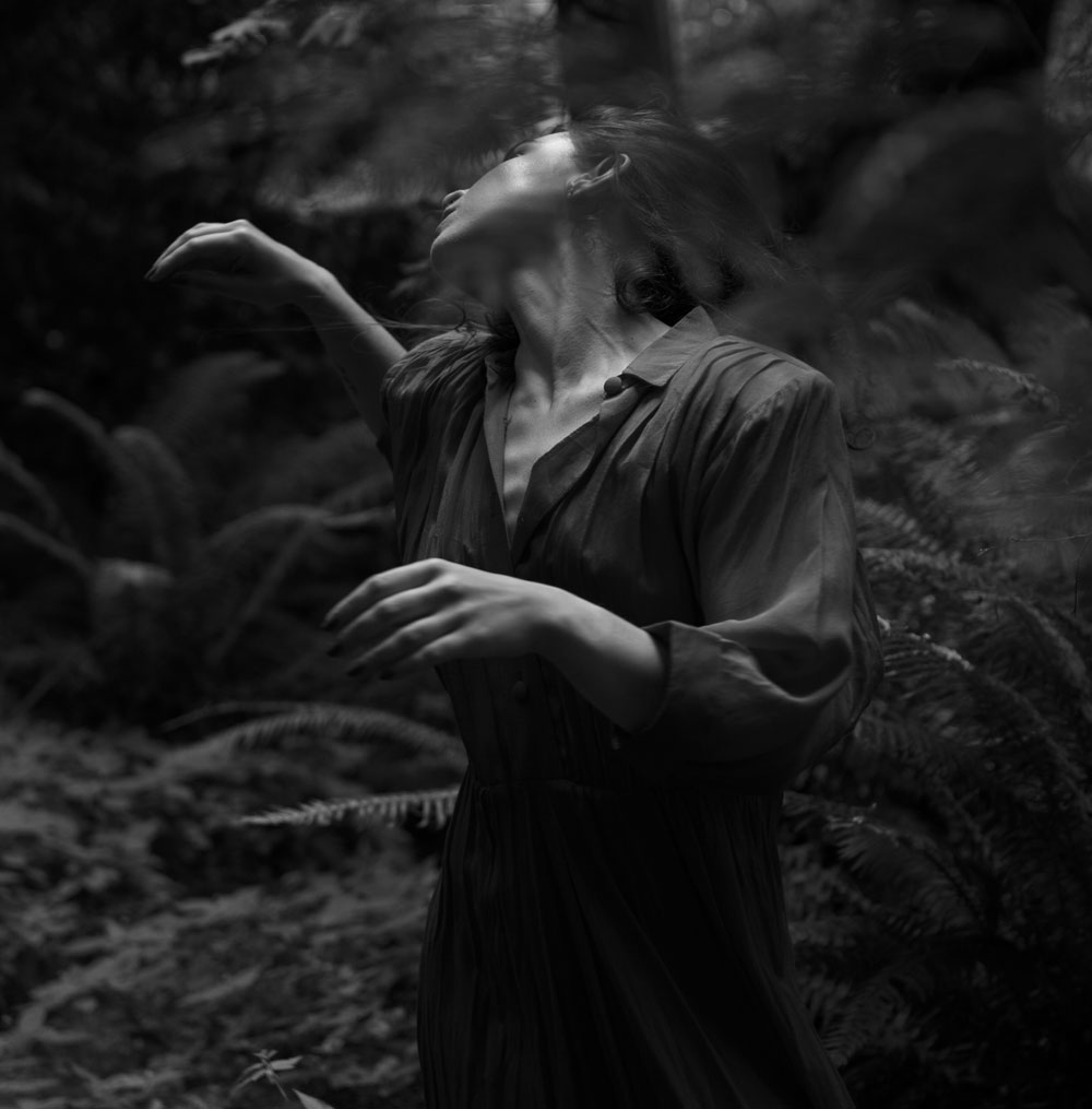 A dance artist poses in a forest. She is wearing a dress and is looking over her right shoulder. Her arms are bent and one is reaching out behind her. The image is in black and white.