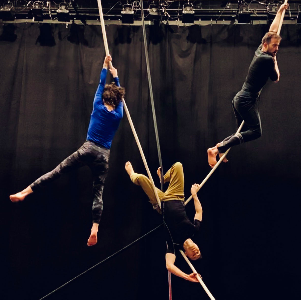 A dance artist hangs from a rope in a black studio while another artist draped on a web of ropes next to her - he is upside down. A third artist climbs a rope up on the right