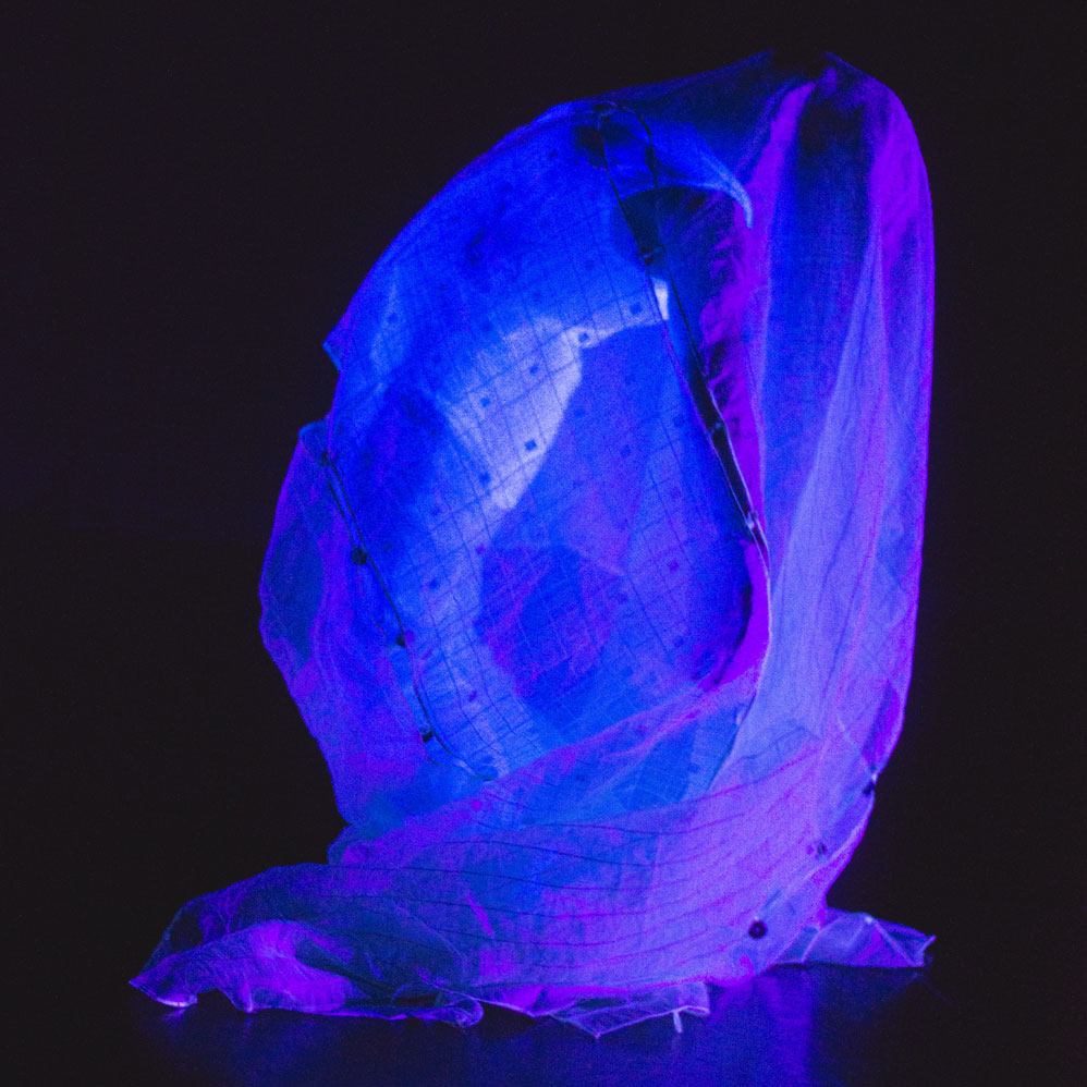 A dance artist arches backwards with their arms stretched out behind them. They are draped in fabric that has a blue tint and are wearing white clothes.