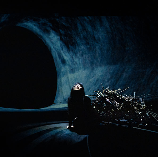 A dance artist sits on a dark stage. Behind them is lit to look like a large cave. The artist is in shadows, and right behind them is a large pile of sticks in a dim spotlight.