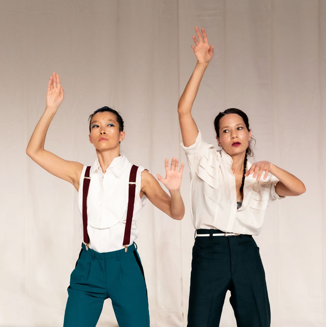 Two dance artists stand in front of white curtains. They are wearing slacks and button up white shirts. The one on the left is wearing suspenders and has her hair pulled back, the other has her hair in two braids. They both have their right arms reached up above them and their left hands bent out in front of them.