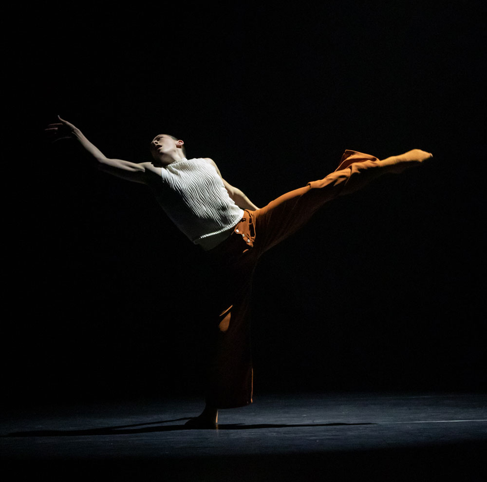 A dance artist poses on a black stage. Their left leg is outstretched as is their right arm and their head is tilted back. They are wearing a sleeveless white sweater and orange pants and socks.