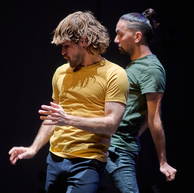 Two dance artists move on a dark stage. One is wearing a yellow t shirt and the other is wearing a green t shirt and has a high brown ponytail.