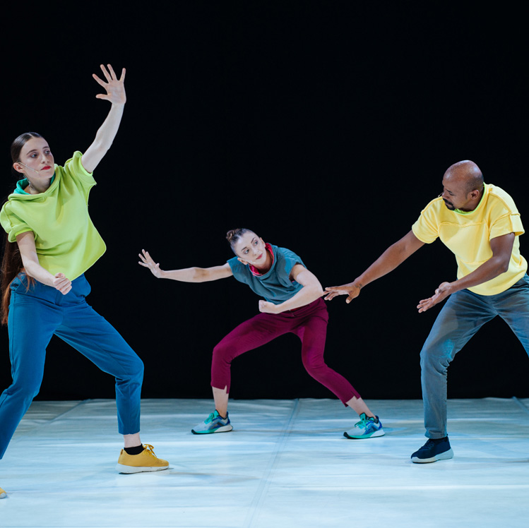 Three dance artists wearing bright clothes pose mid performance. They have glow sticks around their necks and arms and are wearing bright running shoes on the white dance floor.
