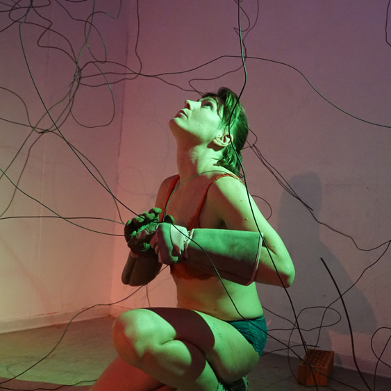 A dance artist kneels down, looking up. They are wearing welding gloves and are surrounded by various wires bent in many shapes.