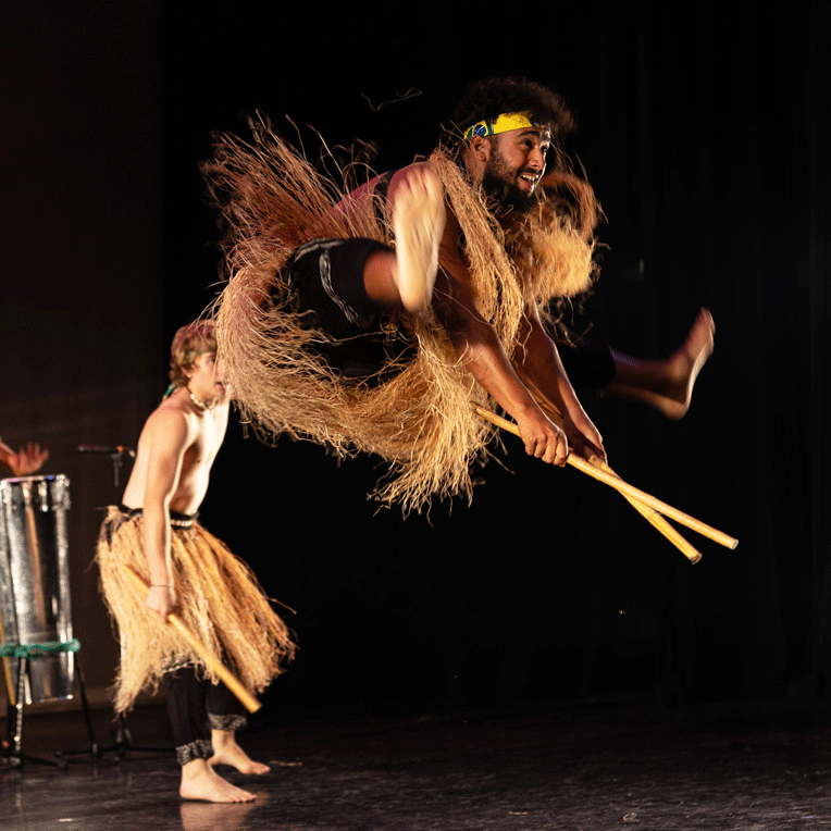 A dance artist jumps high in the air with their legs outstretched. They are wearing a grass skirt. Thee is another dancer standing behind them also wearing a grass skirt.