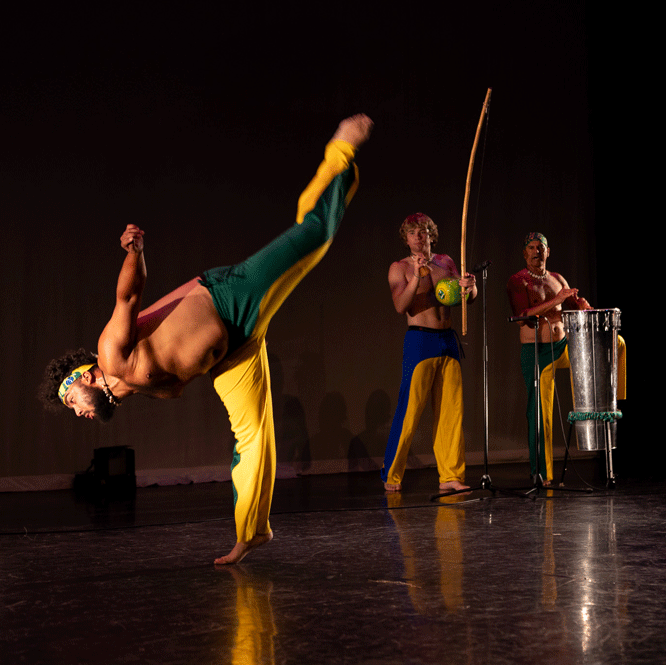A dance artist leans over with one leg very high up behind them. There are two other people standing on the stage playing percussions. They are all wearing bright yellow pants with blue or green paneling.