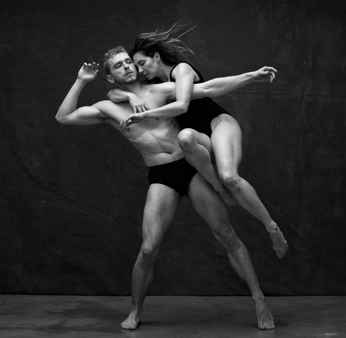 Two dance artists pose. They are wearing minimal clothing and one is hanging off of the other's back with her legs wrapped up around his stomach. This image is in black and white.
