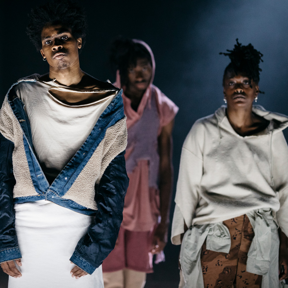 Three dance artists stand looking somber, staring just past the camera. they are wearing a variety of clothing, some of which is torn.