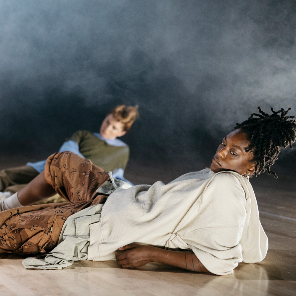 Two dance artists lie on a stage looking just away from the camera. There is one in focus at the front wearing a white hoodie and the other dance artist is out of focus in the background.