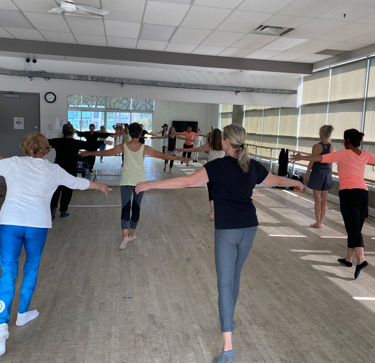 A group of seniors dancing in a bright studio. They are all wearing workout clothing and are facing away from the camera, looking at the teacher.