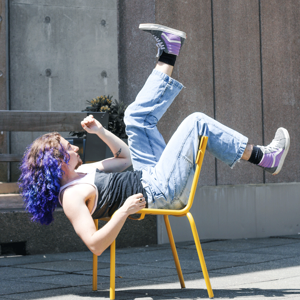 Jay Ginger lies backward on a yellow chair outside. He has one leg over the back of the chair and the other up in the air.
