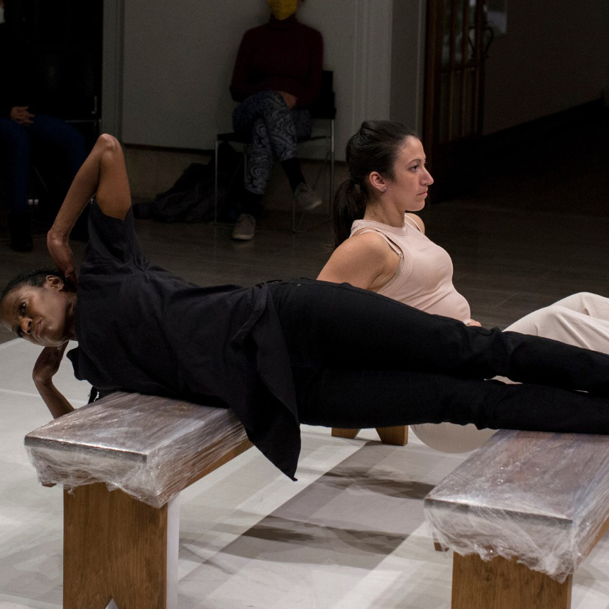 Two dance artists perform on benches covered in plastic. One artists stretches over them on their side, wearing all black with their arms bent up by their head. The other, wearing all white, balances between the two - with most of their body hanging in between.