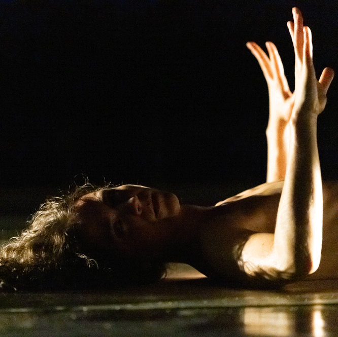 A dance artist lies on a dark stage. They are looking at the camera with their arms bent up. They are topless.