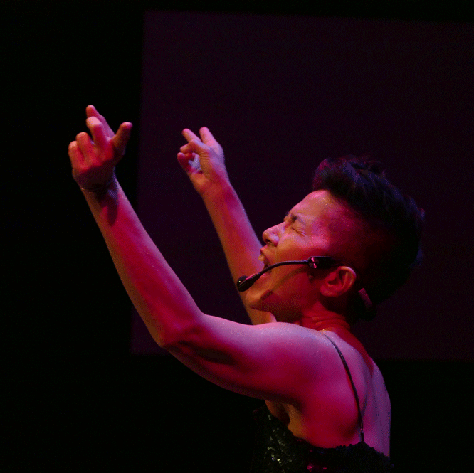A dance artist raises her arms in the arm, mid performance. She appears to be screaming and has a mic on her ear.