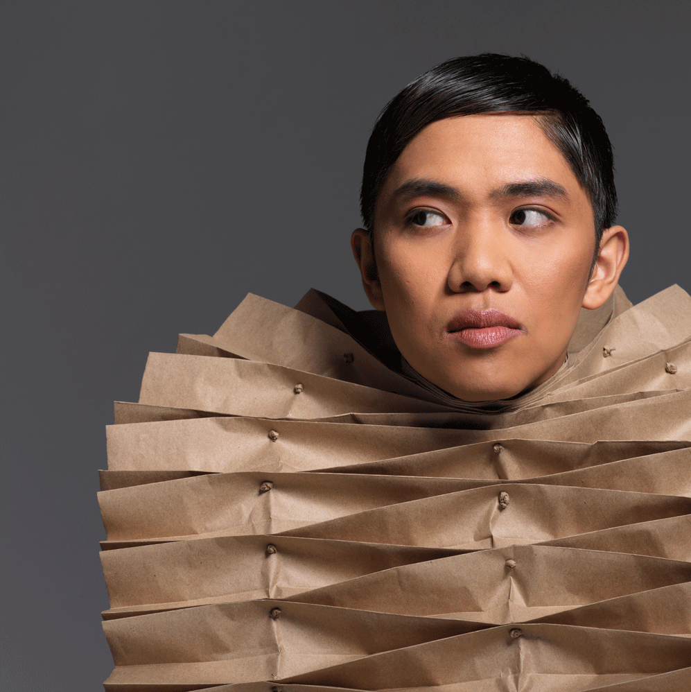 A dance artist poses, looking away from the camera. They are wearing a brown paper structure on their torso - the paper is folding back on itself like a fan.