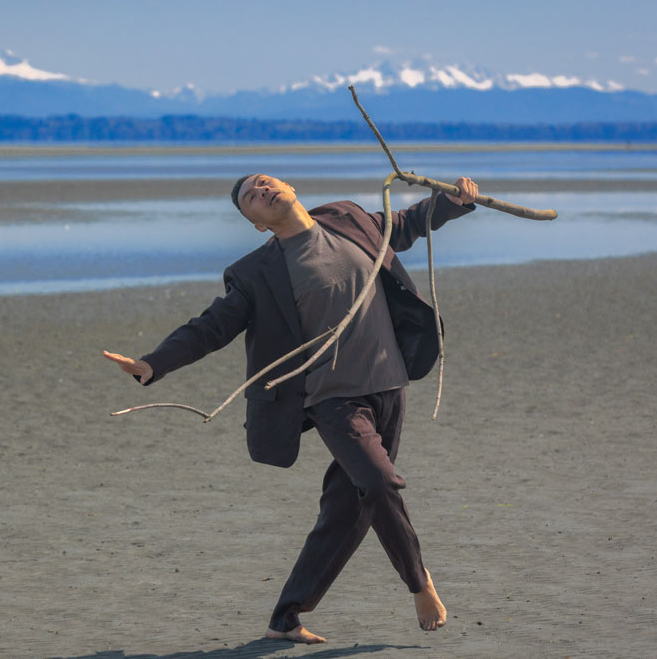 A dance artist stands on a beach in front of water and snowy mountains on a sunny day. He is wearing a dark grey blazer and pants with a light grey shirt under. He has one leg out and his head back and is holding a long multi branched stick.