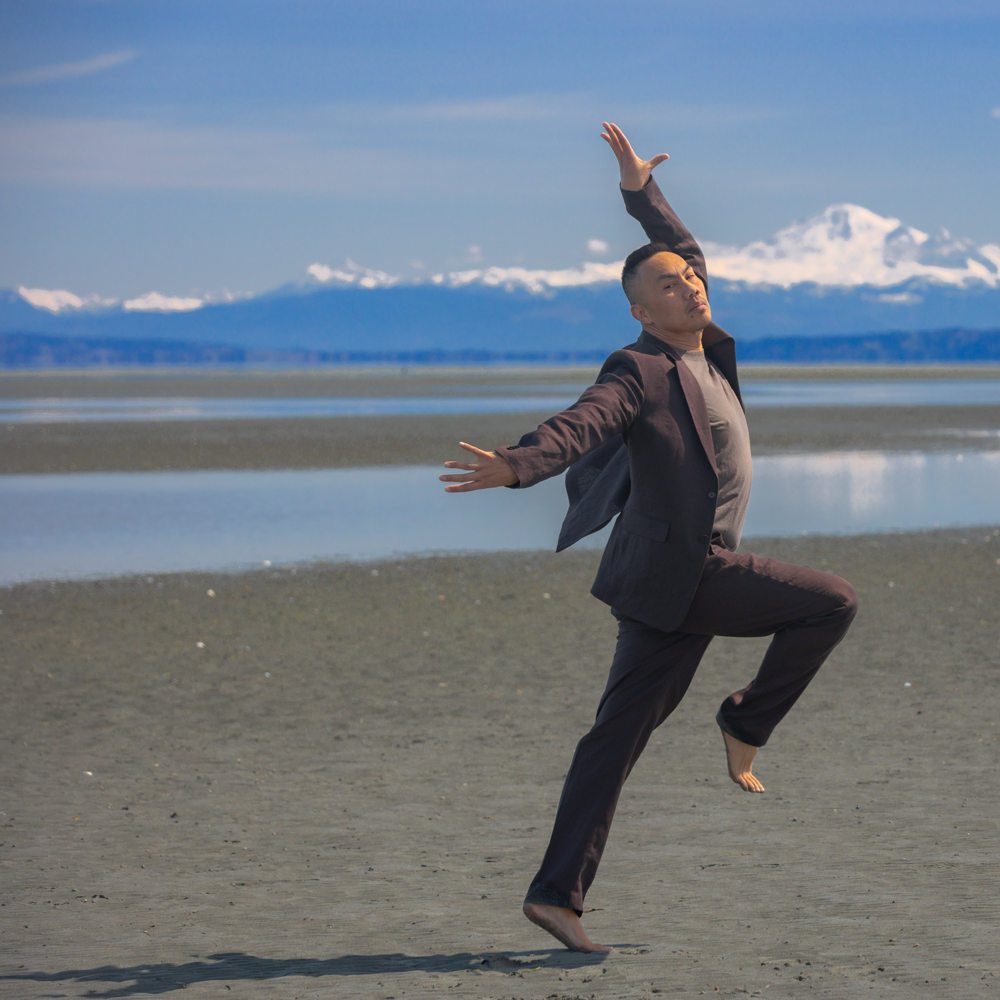 A dance artist stands on a beach in front of water and snowy mountains on a sunny day. He is wearing a dark grey blazer and pants with a light grey shirt under. He has one leg bent up and both arms extended out and up.