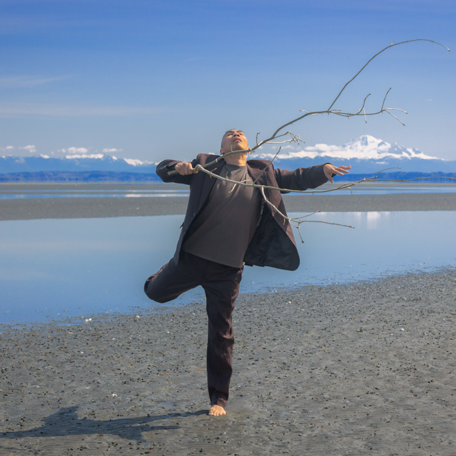 A dance artist stands on a beach in front of water and snowy mountains on a sunny day. He is wearing a dark grey blazer and pants with a light grey shirt under. He has one leg out and his head back and is holding a long multi branched stick.