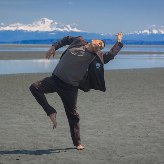 A dance artist stands on a beach in front of water and snowy mountains on a sunny day. He is wearing a dark grey blazer and pants with a light grey shirt under. He has one leg bent up while tilting over with his arms bent out.